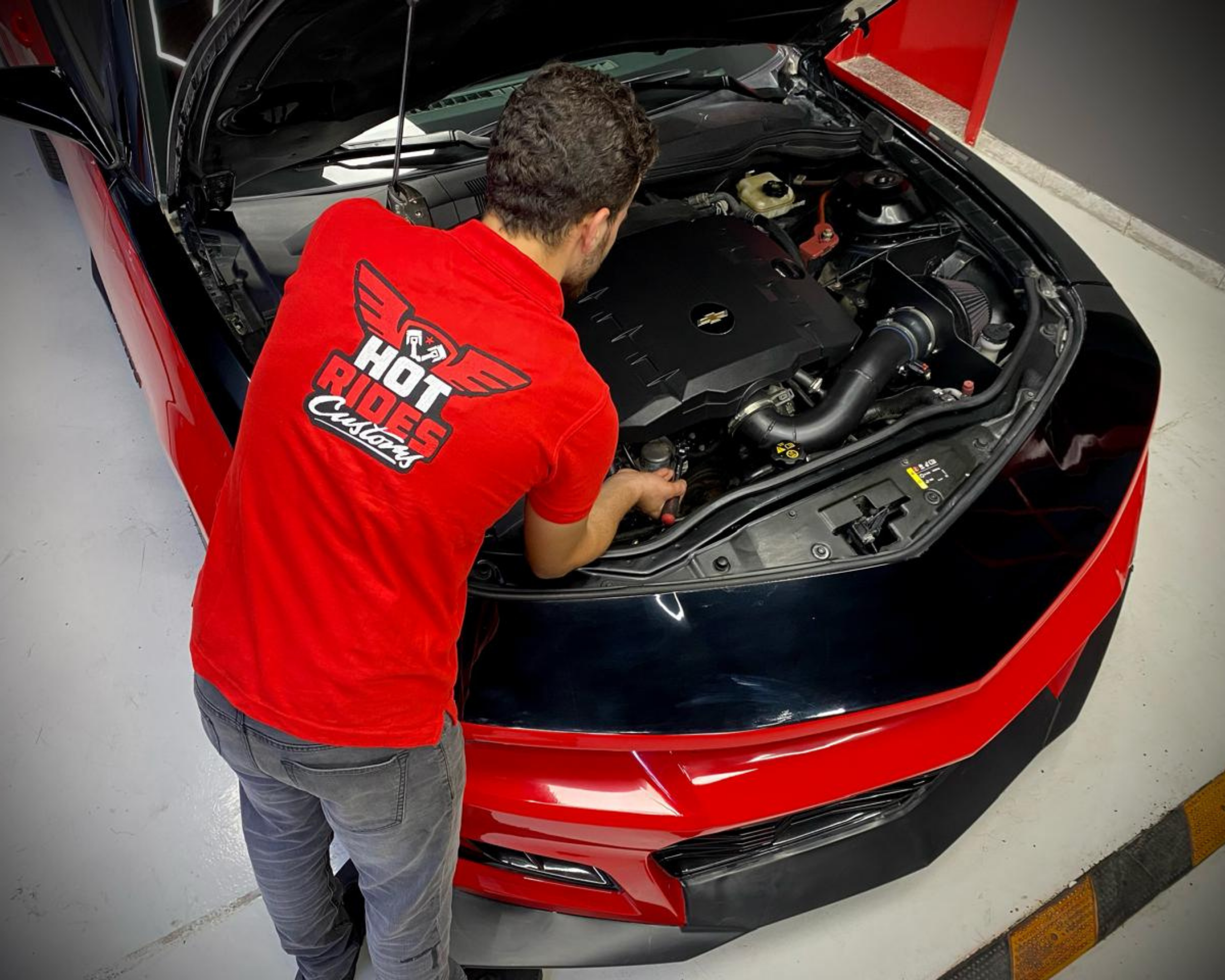 top-down view of mechanic in red servicing red american camaro car doing engine work and general maintenance at Hot Rides Customs Garage in Umm Ramool Al Rashidiya Dubai